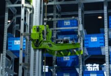Sztuczna inteligencja w logistyce produkcji: mniej przestojów, niższe koszty Zielone ramię robota w zautomatyzowanym magazynie przenosi niebieskie skrzynki